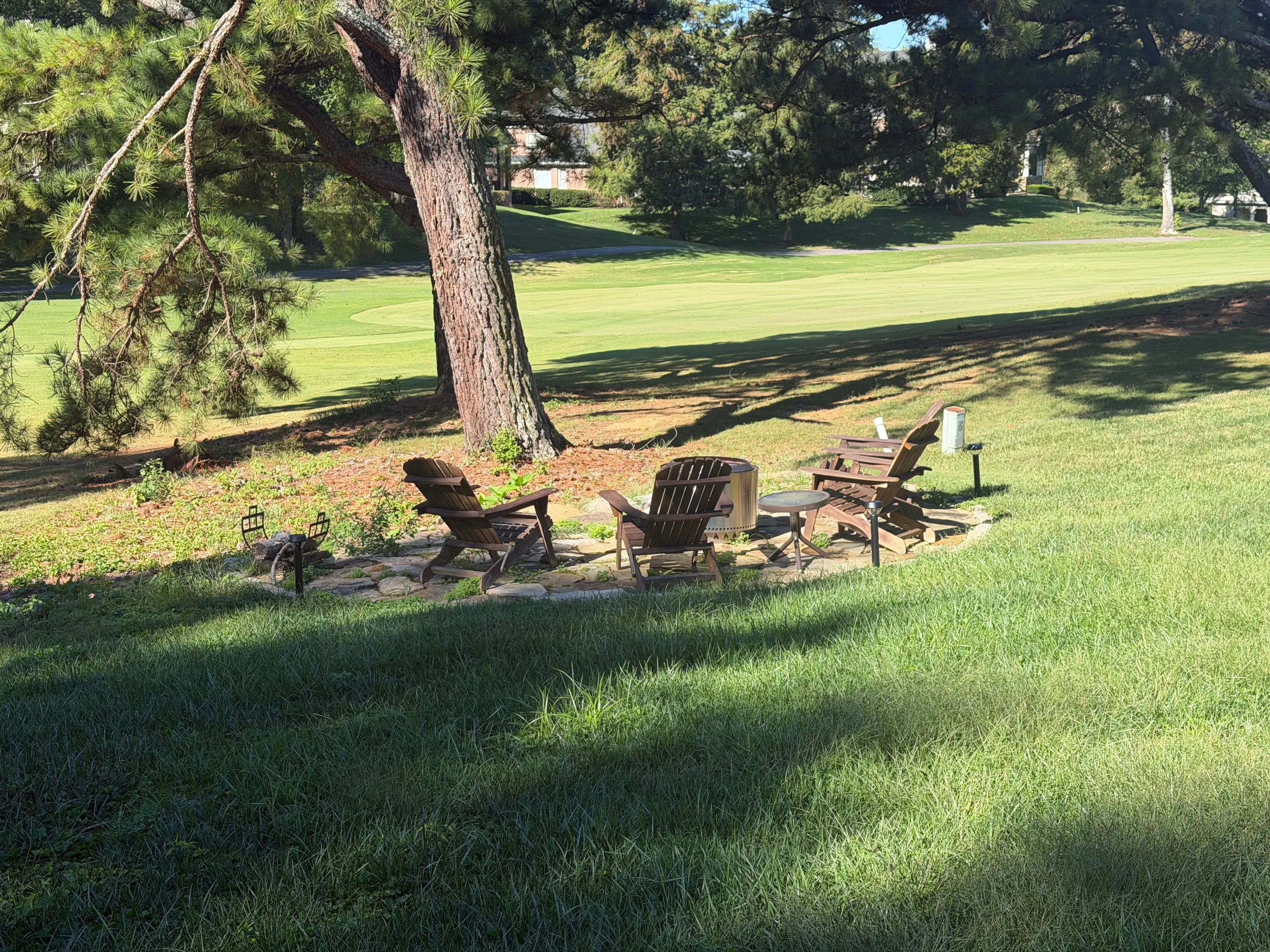 Adirondack chairs around a fire pit on a rock patio under a pine tree, with the golf fairway beyond.