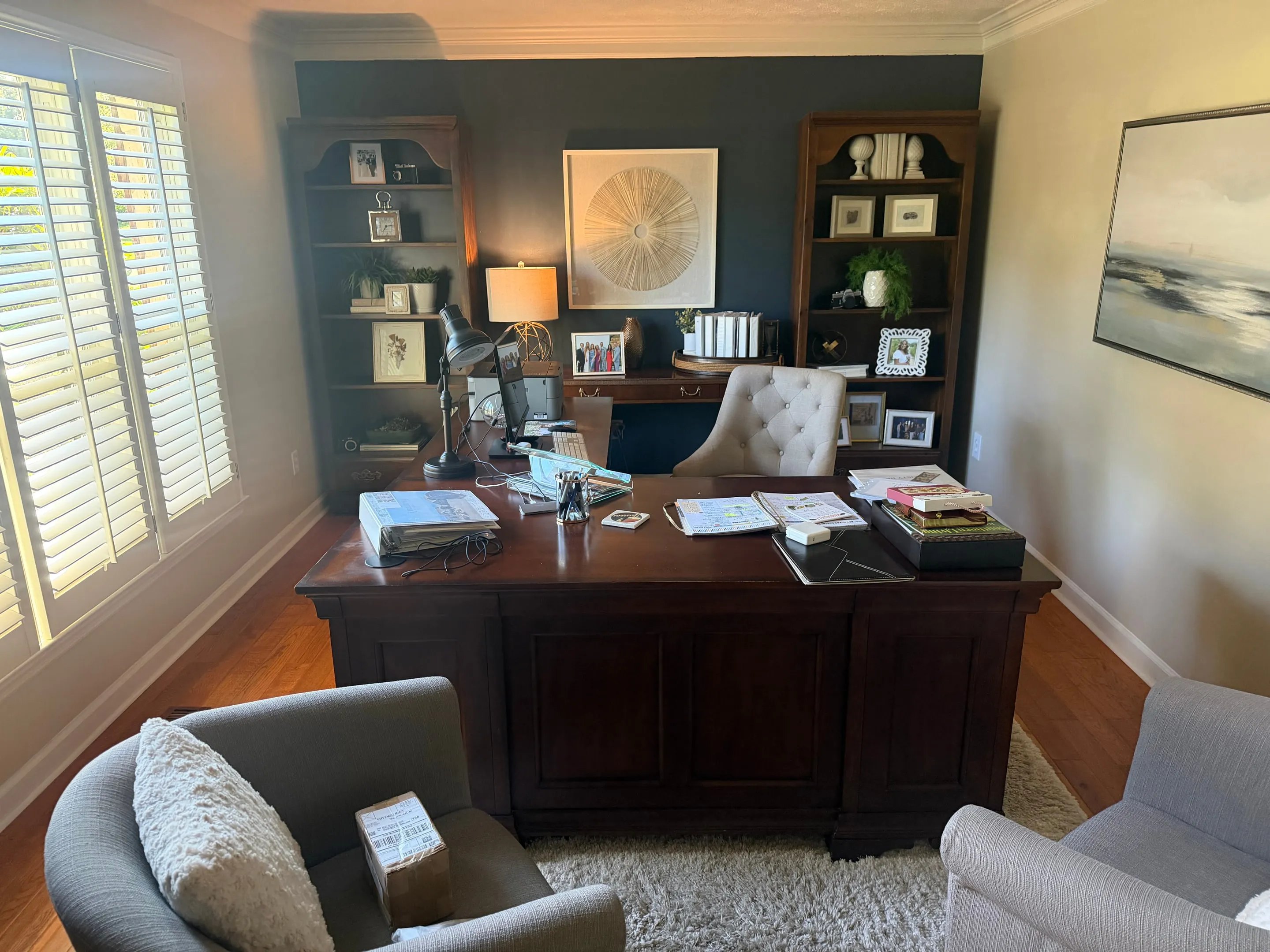 Office with a wood desk, two bookcases against a dark accent wall, and plantation shutters.