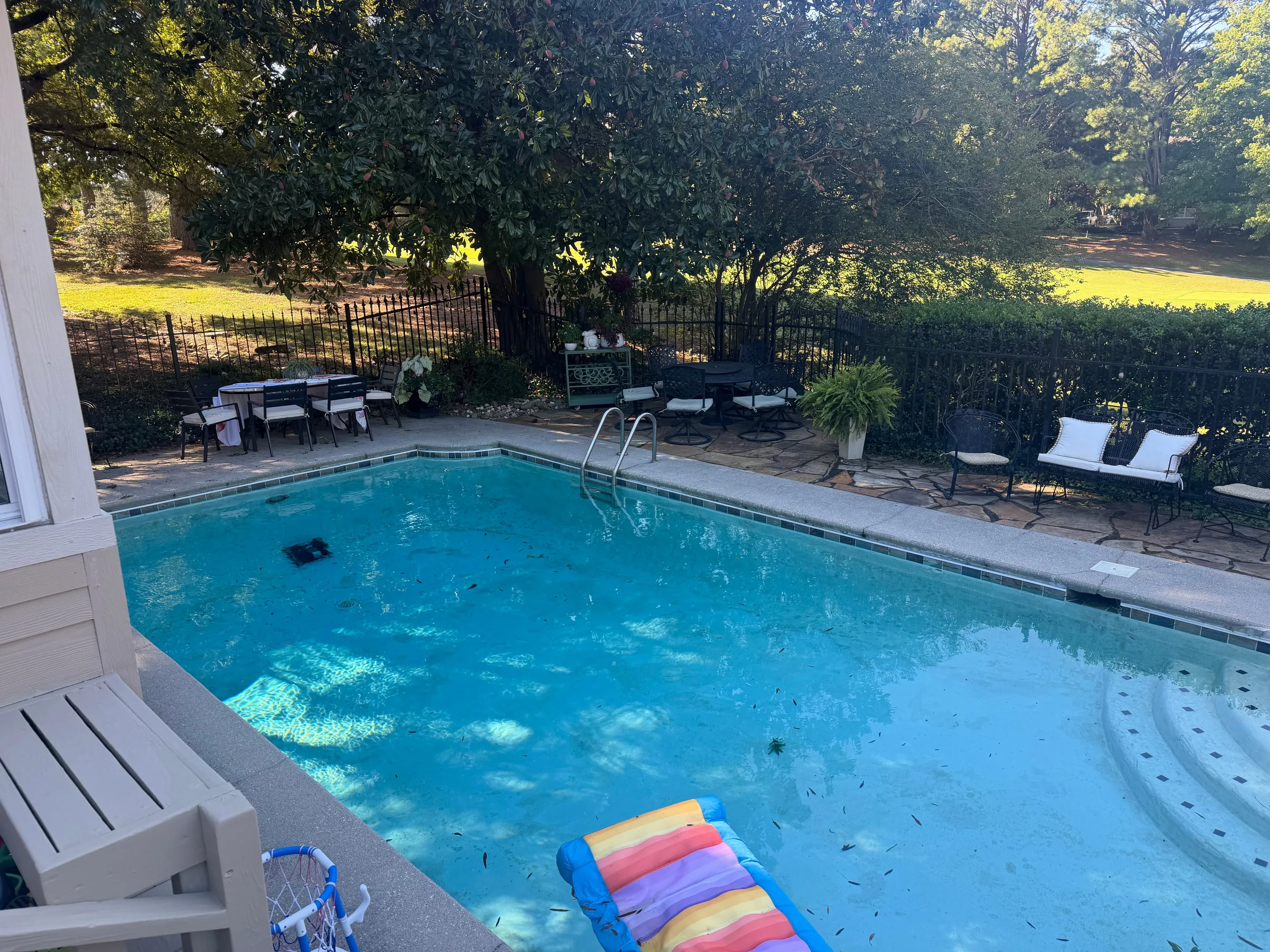 In-ground pool with a concrete deck, flagstone patio, and iron fence under large trees.