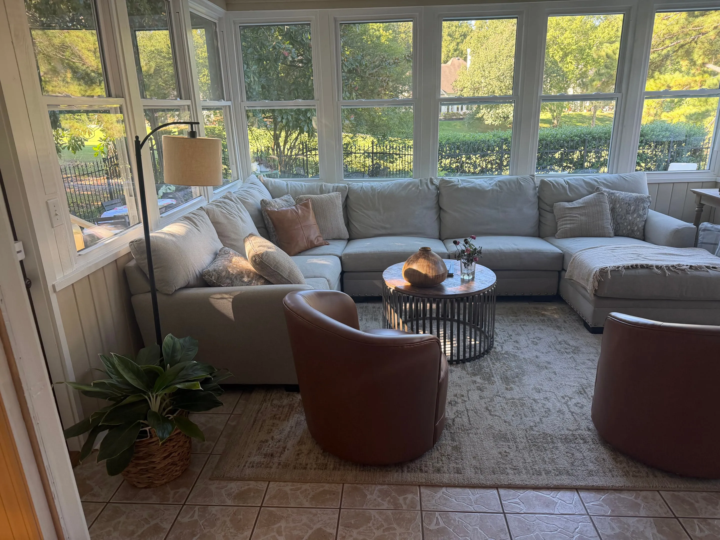 Sunroom with a sectional sofa, tile floor, and windows on three sides looking onto trees.
